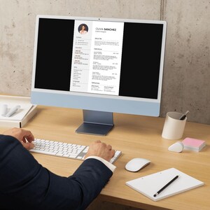 May include: A person working at a desk with a computer displaying a resume. The computer is light blue with a white screen. The resume includes the name Olivia Sanchez. A keyboard, mouse, notepad, and pen are on the desk.