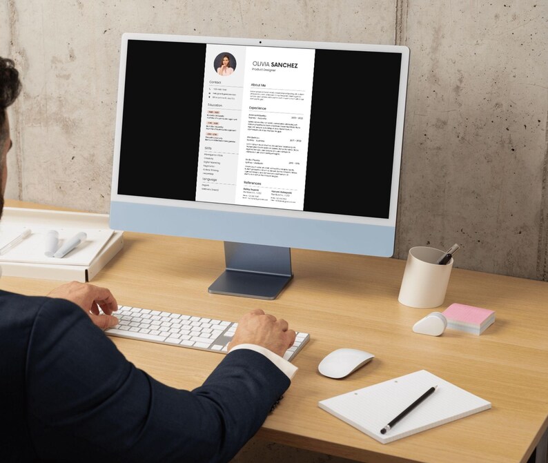 May include: A person working at a desk with a computer displaying a resume. The computer is light blue with a white screen. The resume includes the name Olivia Sanchez. A keyboard, mouse, notepad, and pen are on the desk.