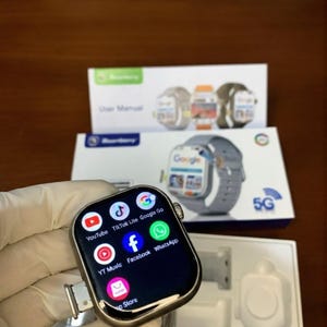 May include: A silver smartwatch with a black screen displaying app icons for YouTube, TikTok, Facebook, and others. The watch is shown with its packaging, which includes a user manual and a picture of the watch. The watch has a gray band.