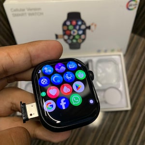 May include: Black smartwatch with a rectangular screen displaying colorful app icons. The watch has a black band and a charging port. The box behind the watch reads "Cellular Version SMART WATCH".