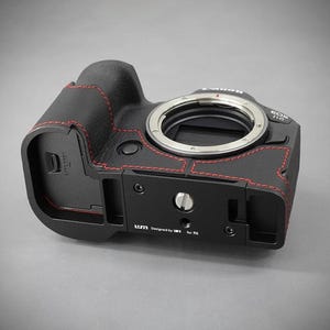 Puede incluir: Cuerpo de c&aacute;mara negro con costuras rojas y montura de lente plateada. La c&aacute;mara tiene una placa base negra con el texto "Designed by LM3 for R6". Se muestra una c&aacute;mara Canon EOS R6.
