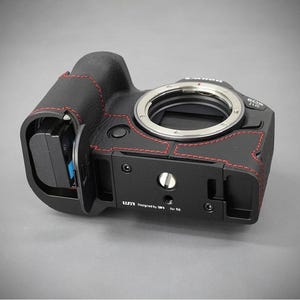 Puede incluir: Cuerpo de c&aacute;mara negro con costuras rojas y montura de lente plateada. La c&aacute;mara tiene una placa base negra con el texto "Designed by UM1 for R6". La c&aacute;mara est&aacute; sobre una superficie gris.