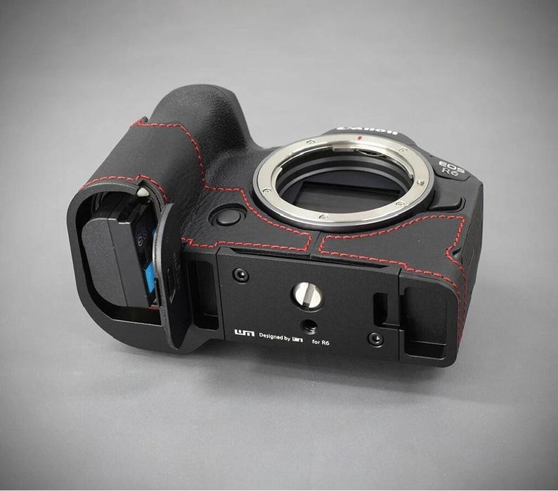 Puede incluir: Cuerpo de c&aacute;mara negro con costuras rojas y montura de lente plateada. La c&aacute;mara tiene una placa base negra con el texto "Designed by UM1 for R6". La c&aacute;mara est&aacute; sobre una superficie gris.