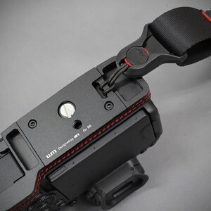 Puede incluir: C&aacute;mara negra con una correa negra y roja adjunta. La c&aacute;mara tiene una base de metal con costuras rojas y el texto "Designed by UM for R6". La correa tiene una hebilla negra con un acento rojo.