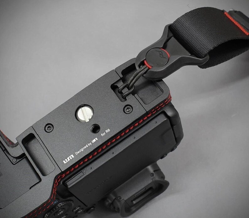 Puede incluir: C&aacute;mara negra con una correa negra y roja adjunta. La c&aacute;mara tiene una base de metal con costuras rojas y el texto "Designed by UM for R6". La correa tiene una hebilla negra con un acento rojo.