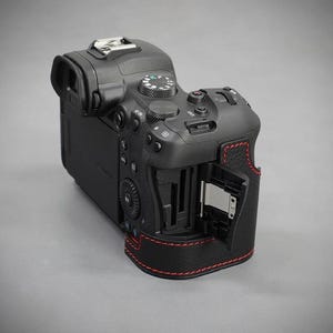 Puede incluir: C&aacute;mara digital negra con costuras rojas. La c&aacute;mara tiene m&uacute;ltiples botones y diales, un visor y una ranura para tarjetas abierta. Sobre un fondo gris.