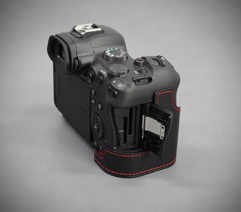 Puede incluir: C&aacute;mara digital negra con costuras rojas. La c&aacute;mara tiene m&uacute;ltiples botones y diales, un visor y una ranura para tarjetas abierta. Sobre un fondo gris.