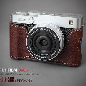 Könnte beinhalten: Eine silberne Fujifilm X-E5 Kamera mit einer braunen Leder-Halbhülle. Das Kameraobjektiv ist schwarz mit weißer Schrift. Der Text auf der Hülle lautet "FJ-XE5BR (BROWN)" und "FUJIFILM X-E5".