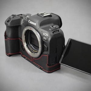 Puede incluir: C&aacute;mara Canon EOS R6 negra con funda de cuero y costuras rojas. La c&aacute;mara tiene una montura de lente, una pantalla abatible y el logotipo de Canon. La funda envuelve el cuerpo de la c&aacute;mara.