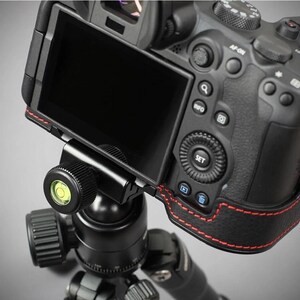 Puede incluir: Una c&aacute;mara digital negra montada en una r&oacute;tula de tr&iacute;pode negra. La c&aacute;mara tiene una pantalla negra y costuras rojas en el lateral. La r&oacute;tula del tr&iacute;pode tiene un nivel con una burbuja verde.