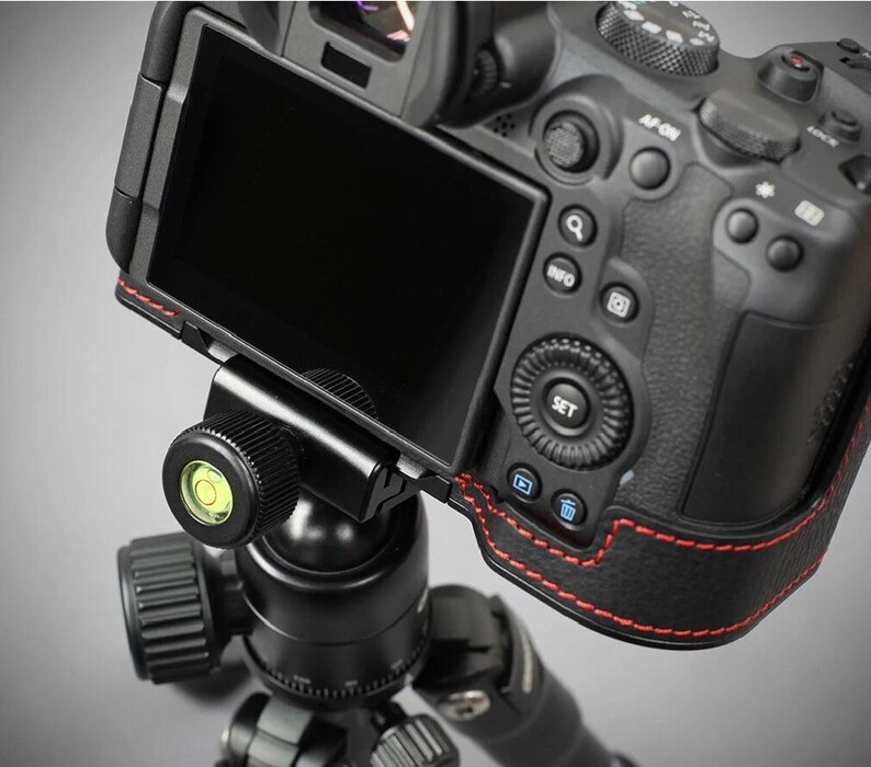 Puede incluir: Una c&aacute;mara digital negra montada en una r&oacute;tula de tr&iacute;pode negra. La c&aacute;mara tiene una pantalla negra y costuras rojas en el lateral. La r&oacute;tula del tr&iacute;pode tiene un nivel con una burbuja verde.