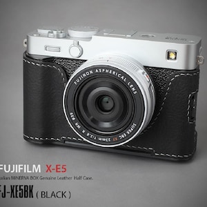 Könnte beinhalten: Eine silberne und schwarze Fujifilm X-E5 Kamera mit einer schwarzen Leder-Halbhülle. Das Kameraobjektiv ist schwarz mit weißer Schrift. Der Text auf dem Bild lautet "FUJIFILM X-E5" und "FJ-XE5BK (BLACK)".