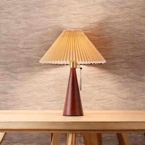May include: A wooden table lamp with a pleated lampshade. The lamp has a dark brown, conical base and a light tan lampshade. A small metal pull chain hangs from the lamp. The lamp is on a wooden table.