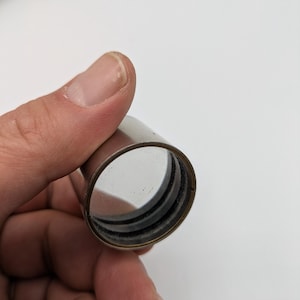 Puede incluir: Un tubo de metal plateado con una lente de vidrio transparente en su interior. El tubo tiene un anillo de goma negro alrededor del borde de la lente.