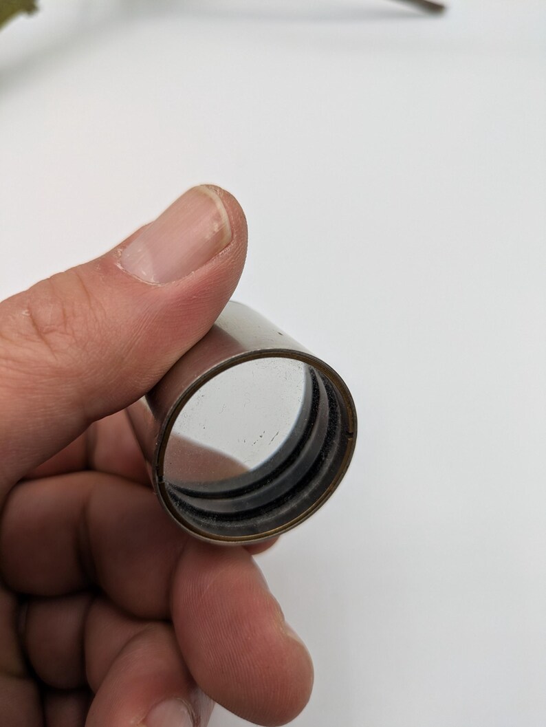 Puede incluir: Un tubo de metal plateado con una lente de vidrio transparente en su interior. El tubo tiene un anillo de goma negro alrededor del borde de la lente.