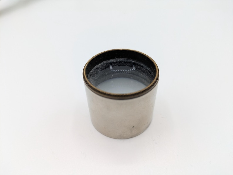 Puede incluir: Una tapa de lente de plata y lat&oacute;n con un anillo de goma negro.