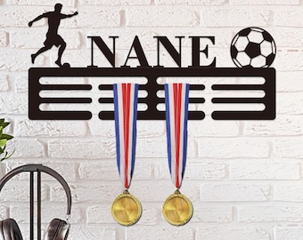 May include: A black metal soccer-themed medal holder with the name "NANE" cut out. The holder features a soccer player silhouette, a soccer ball, and three horizontal bars for hanging medals. Two gold medals with red, white, and blue ribbons are displayed.