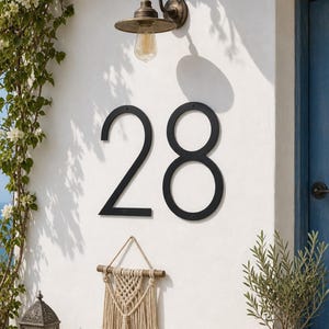 May include: Large black metal house numbers 28 mounted on a white wall. The numbers feature a clean, sans-serif font with visible mounting screws. Above the numbers is a bronze-toned wall lamp, and below is a decorative beige macrame wall hanging.