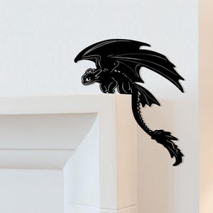 May include: A black dragon silhouette with outstretched wings and a long tail. The dragon is perched on a white surface, with its head and body visible. The wings are spread wide, and the tail curves downward.
