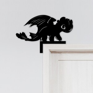 May include: Black silhouette of a dragon character, with large wings and a friendly expression. The dragon is perched on a black ledge, designed to be placed on a door frame. The design is a decorative element.