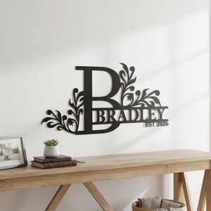 May include: A black metal wall sign featuring a large capital letter B with decorative floral scrollwork extending from the sides. The name BRADLEY is integrated into the design, accompanied by the text EST 2026 in a clean, sans-serif font.