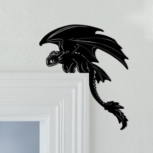 May include: Black dragon-shaped wall decor. The dragon has large wings, a long tail, and is perched on a white door frame. The dragon is a silhouette, with detailed features. This decorative piece is suitable for a child's room or a fantasy-themed space.
