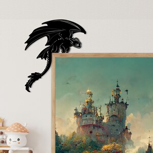 May include: A black dragon-shaped wall decor piece. The dragon is perched on the corner of a framed picture with a fantasy castle scene. The dragon has large wings and a long tail. The artwork is a decorative accent for a room.
