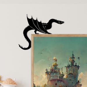 May include: A black metal dragon silhouette decorates the corner of a framed artwork. The dragon has outstretched wings and a long tail. The artwork features a fantasy castle scene with a teal sky.