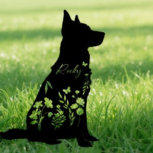 May include: A black silhouette of a dog sitting in profile, filled with green floral and butterfly designs. The name "Rocky" is written in a cursive font on the dog's side. The background is a blurred green field.