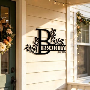 May include: A black metal wall sign featuring a large capital letter B with decorative floral scrollwork on the left and right sides. The name BRADLEY is centered in the middle, with the text EST 2026 positioned directly underneath the name.