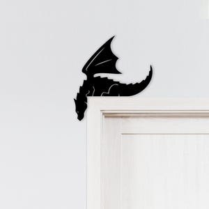 May include: A black dragon silhouette decorates the corner of a white door. The dragon has outstretched wings and a long tail, creating a fantasy-themed accent. The dragon is a decorative element.