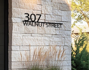 Könnte beinhalten: Schwarzes Metall-Adressschild mit den Zahlen "307" über den Worten "WALNUT STREET". Das Schild ist an einer hellfarbigen Steinmauer angebracht. Ziergräser und Grünpflanzen befinden sich im Vordergrund.