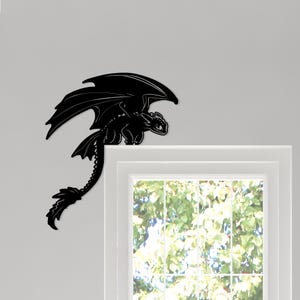 May include: A black dragon wall decor piece. The dragon has large wings and a long tail, perched on a white window frame. The dragon is a silhouette, with detailed features. The background is a light gray wall and a window with a view of green foliage.