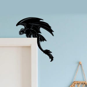 May include: Black dragon wall decor. The dragon is perched on a white door frame, with its wings spread and tail hanging down. The dragon is a silhouette, with intricate details. The background is a light blue wall.