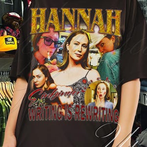May include: A black t-shirt featuring the name "HANNAH" in gold lettering above a collage of images. The shirt also includes the text "Girlboss Writing is Rewriting" in a stylized font. The model is wearing large hoop earrings.