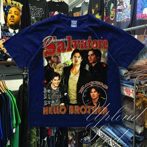 Pu&ograve; includere: T-shirt blu navy con una grafica di diversi uomini e la scritta "Damon Salvatore" in rosso e oro. La maglietta include anche le frasi "Best Night Ever" e "Hello Brother" in stile vintage.