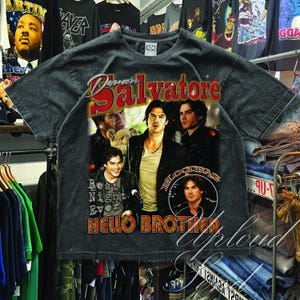 Pu&ograve; includere: T-shirt grigio scuro con un collage di immagini e la scritta "Damon Salvatore" in rosso e bianco, e "Hello Brother" in arancione. La maglietta ha un aspetto vintage e usurato.