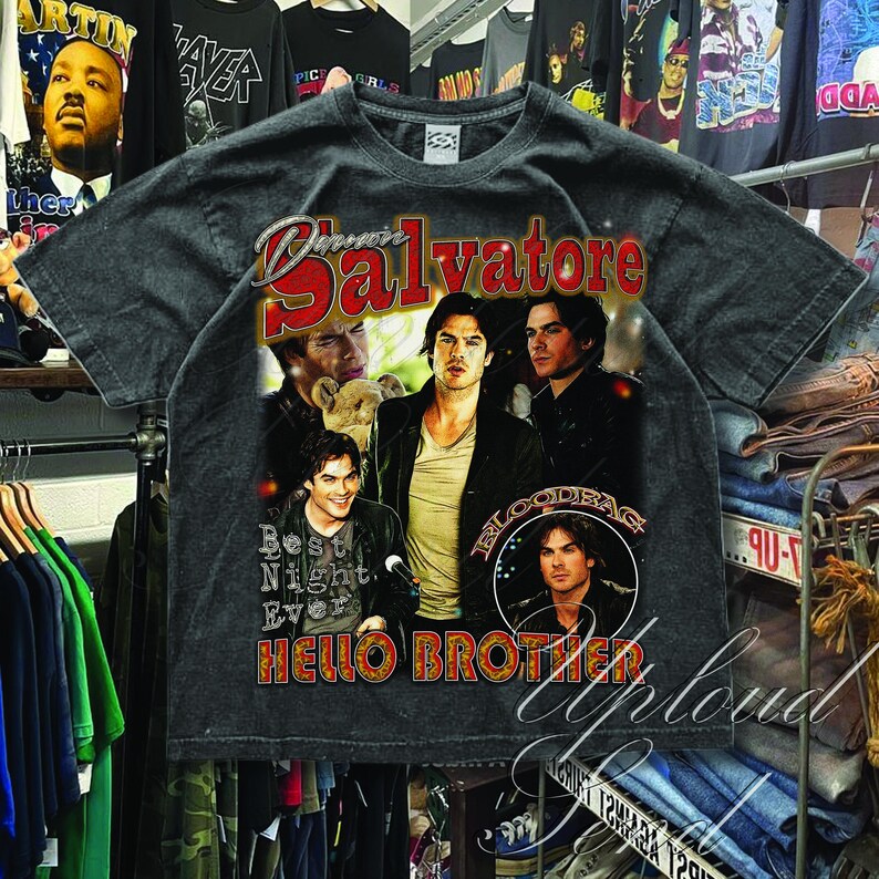 Pu&ograve; includere: T-shirt grigio scuro con un collage di immagini e la scritta "Damon Salvatore" in rosso e bianco, e "Hello Brother" in arancione. La maglietta ha un aspetto vintage e usurato.