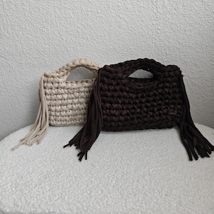 May include: Two crochet handbags in the image. One is beige, the other is dark brown. Both have a rectangular shape with a handle and fringe detail. The bags are sitting on a white, textured surface.