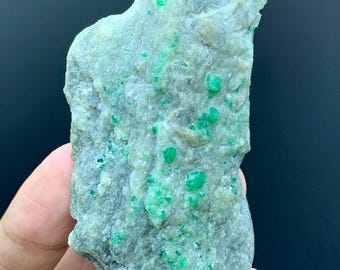 May include: A rough-hewn, irregularly shaped stone with a gray-green hue. The surface is speckled with vibrant, emerald-green crystals. The stone is held in a hand, showcasing its natural texture and color variations. This raw gemstone is a unique mineral specimen.