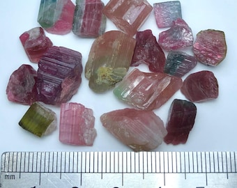 May include: A collection of raw tourmaline crystals in various colors, including pink, green, and blue. The crystals are of different sizes and shapes, with visible striations and natural formations. A ruler is placed next to the crystals for scale.