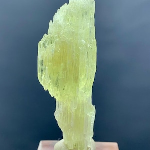 Könnte beinhalten: Ein durchscheinendes, hellgrünes Mineralexemplar. Die Kristallstruktur ist säulenförmig, mit vertikalen Streifen. Der Sockel ist hellgrau, und das Mineral ist auf einem transparenten, rechteckigen Sockel montiert. Der Hintergrund ist dunkelblau-grau.