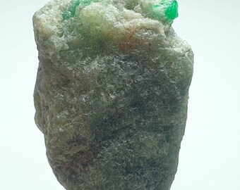 May include: A rough, uncut emerald specimen. The stone has a dark gray-green base with bright green emerald crystals protruding from the top. The specimen is displayed on a small, light-colored stone base against a white background.
