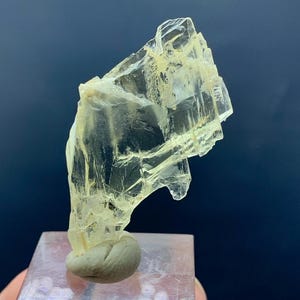 Könnte beinhalten: Ein durchscheinendes, hellgelbes Mineralpräparat mit geschichteter, kristalliner Struktur. Das Mineral ist auf einem transparenten, quadratischen Sockel mit einem kleinen, abgerundeten, grauen Stein montiert. Der Hintergrund ist dunkelblau.