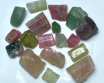 May include: A collection of raw tourmaline crystals in various colors, including green, pink, and peach. The gemstones are irregularly shaped and display natural textures. The crystals are scattered across a white surface, showcasing their diverse hues and forms.
