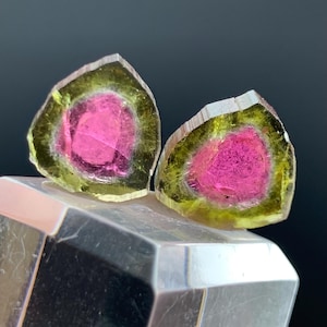 May include: Two watermelon tourmaline slices, showcasing a vibrant pink center surrounded by a green and yellow outer layer. The gemstones are displayed on a clear, faceted crystal base, set against a dark background. The tourmaline crystals are cut to reveal their internal color variations.