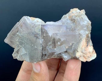 May include: A raw, natural fluorite crystal cluster. The mineral features a mix of gray, purple, and clear crystalline structures. The rough edges and varied textures give it a unique, organic appearance. The crystal is held in a hand against a dark background.