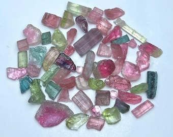 May include: A collection of raw tourmaline crystals in various colors, including pink, green, blue, and purple. The gemstones are irregularly shaped and vary in size, with a mix of translucent and opaque textures.