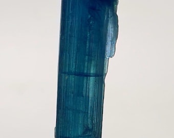 Puede incluir: Un vibrante espécimen de cristal azul alargado. El cristal tiene una estructura columnar con estrías visibles y una superficie ligeramente rugosa y texturizada. El color es un azul profundo y saturado, con variaciones de tono. El cristal se muestra sobre un fondo blanco liso.