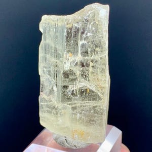 Könnte beinhalten: Ein durchscheinendes, hellgelbes Kristallexemplar. Das Mineral hat eine rechteckige Form mit sichtbaren inneren Strukturen und ist auf einem klaren, geometrischen Präsentationsständer montiert. Der Hintergrund ist dunkelblau.